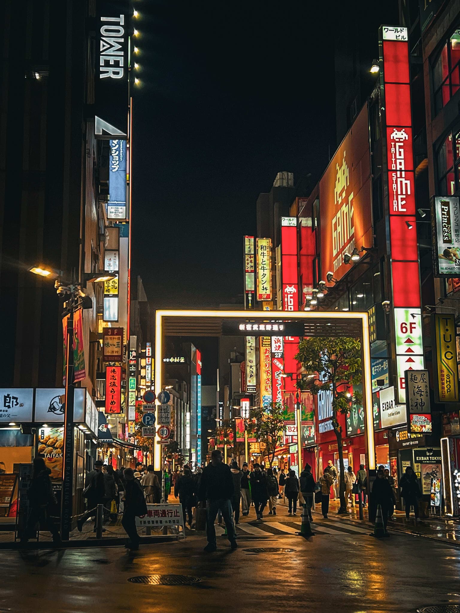 Pedestrians on a rain-slicked Shinjuku street at night, beneath tall red neon signboards and a lit gateway arch